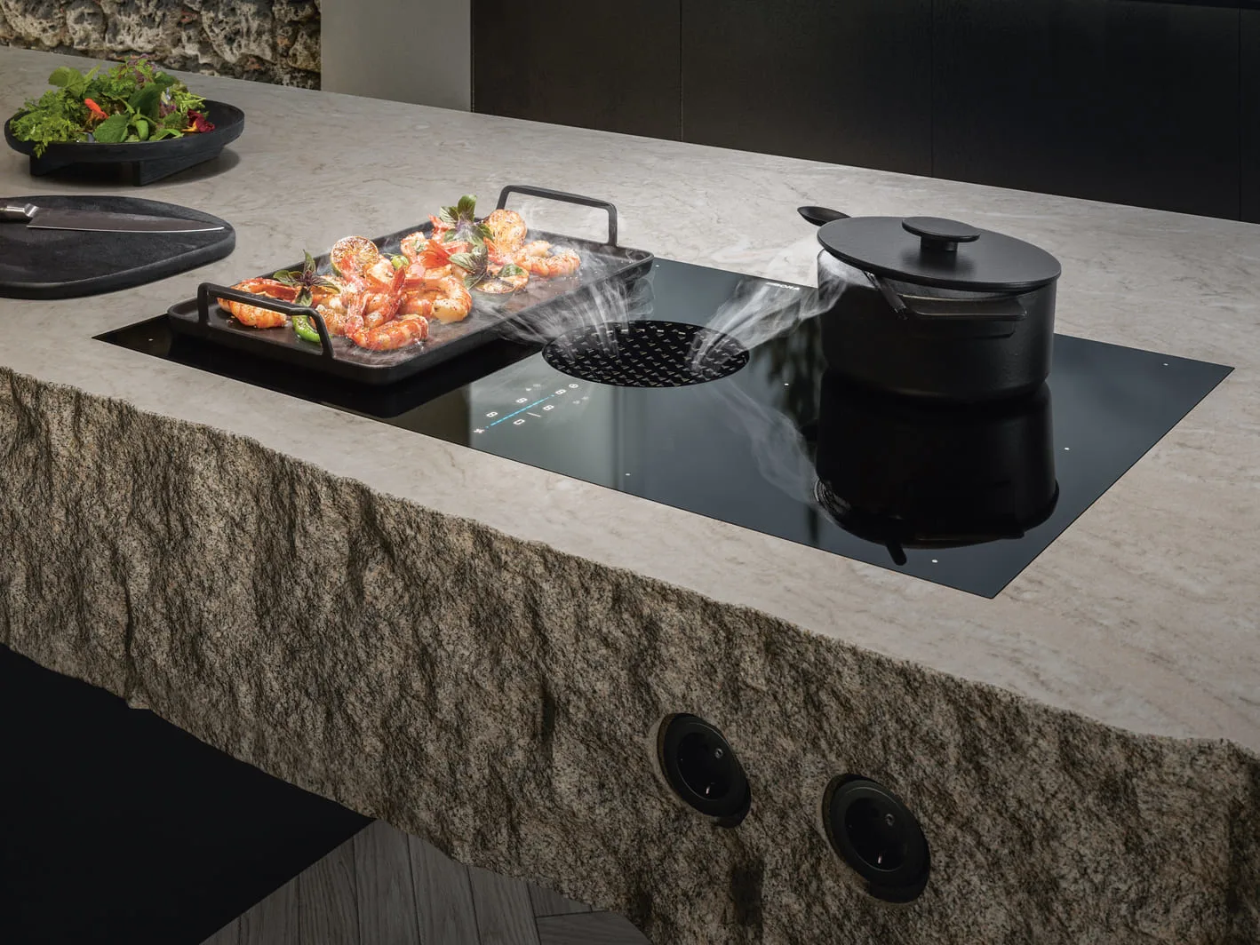 2 BORA X Pure ploča za kuvanje sa integrisanim aspiratorom, grilom i crnim posuđem na radnoj ploči od rustičnog kamena, u toku pripreme hrane.