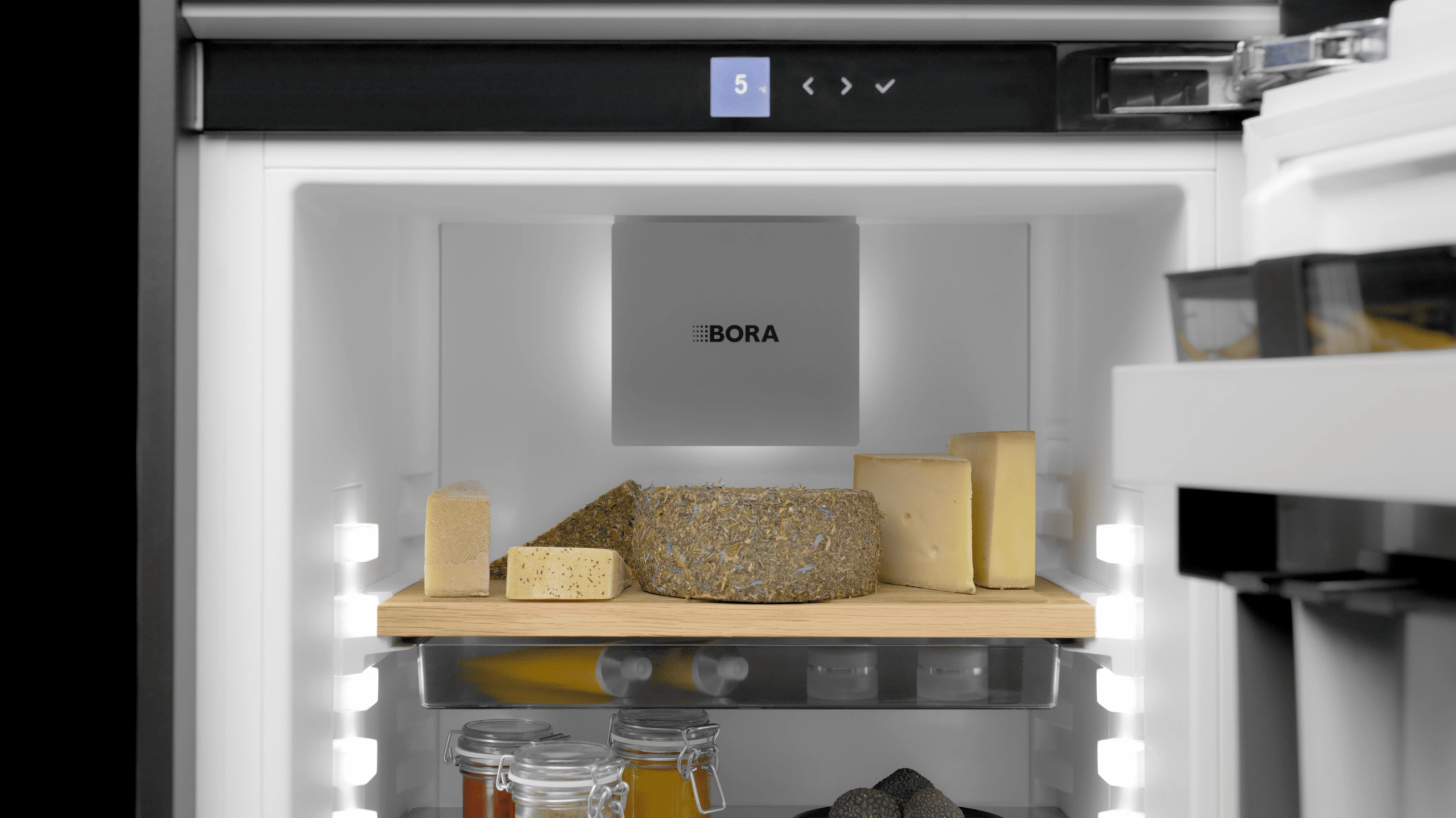4 Unutrašnjost BORA Cool frižidera sa različitim vrstama sireva na drvenoj polici i BORA filterom sa logotipom na zadnjem zidu.