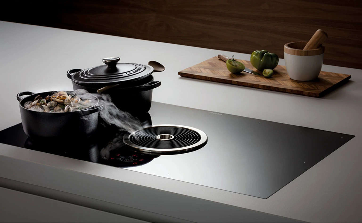 8 BORA Basic aspiraciona ploča za kuvanje, pogled sa strane, crna staklena površina i lonci od livenog gvožđa sa parom tokom kuvanja.
