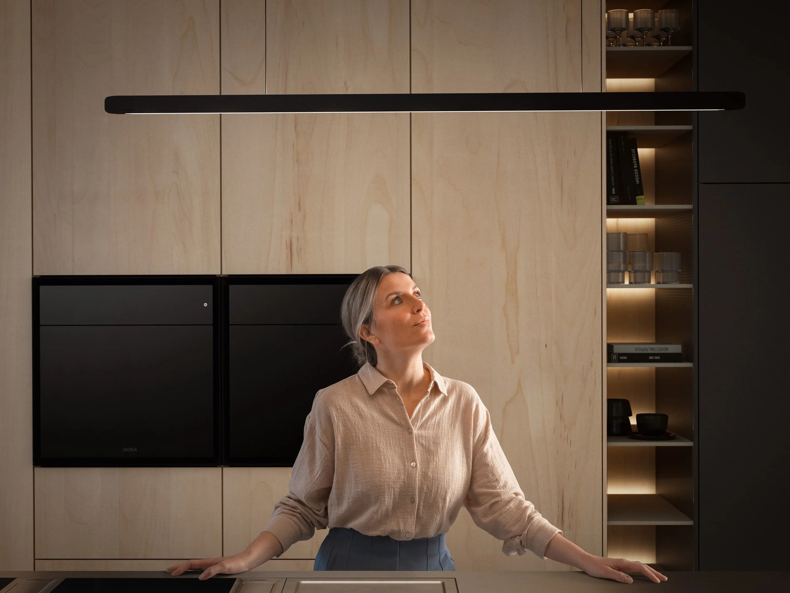 o8ikj8x79dhsvbtrla03 Žena posmatra BORA STARS Horizon lampu u minimalističkoj kuhinji od svetlog drveta, fokus na eleganciji i preciznom dizajnu svetla.